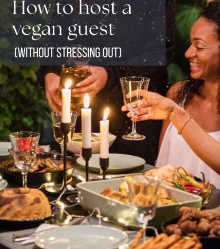 Two smiling women share a toast at a warmly lit dinner table decorated with candles and vegan dishes, illustrating a cozy holiday gathering. Text overlay reads “How to host a vegan guest (without stressing out).”